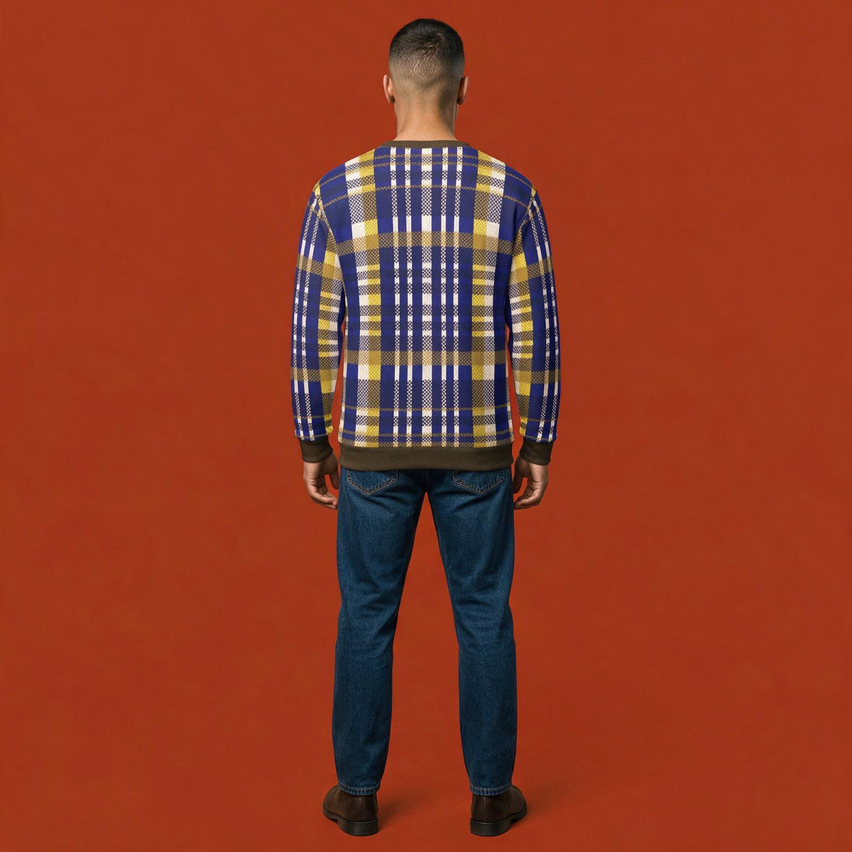 A man viewed from behind wearing a blue, yellow, and white plaid sweater and blue jeans, standing against a solid orange-red background.