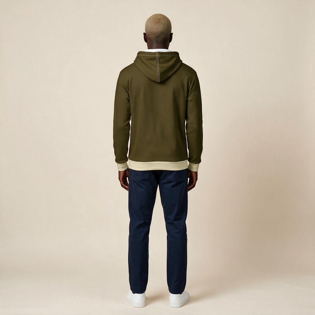 A man viewed from the back wearing an olive green hoodie with beige ribbed trim, navy pants, and white sneakers, standing against a plain beige studio background.
