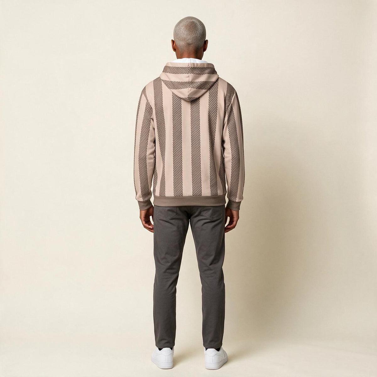 A man viewed from behind wearing a beige and brown vertically striped hoodie with a textured knit pattern, dark gray pants, and white sneakers, standing against a plain beige studio background.