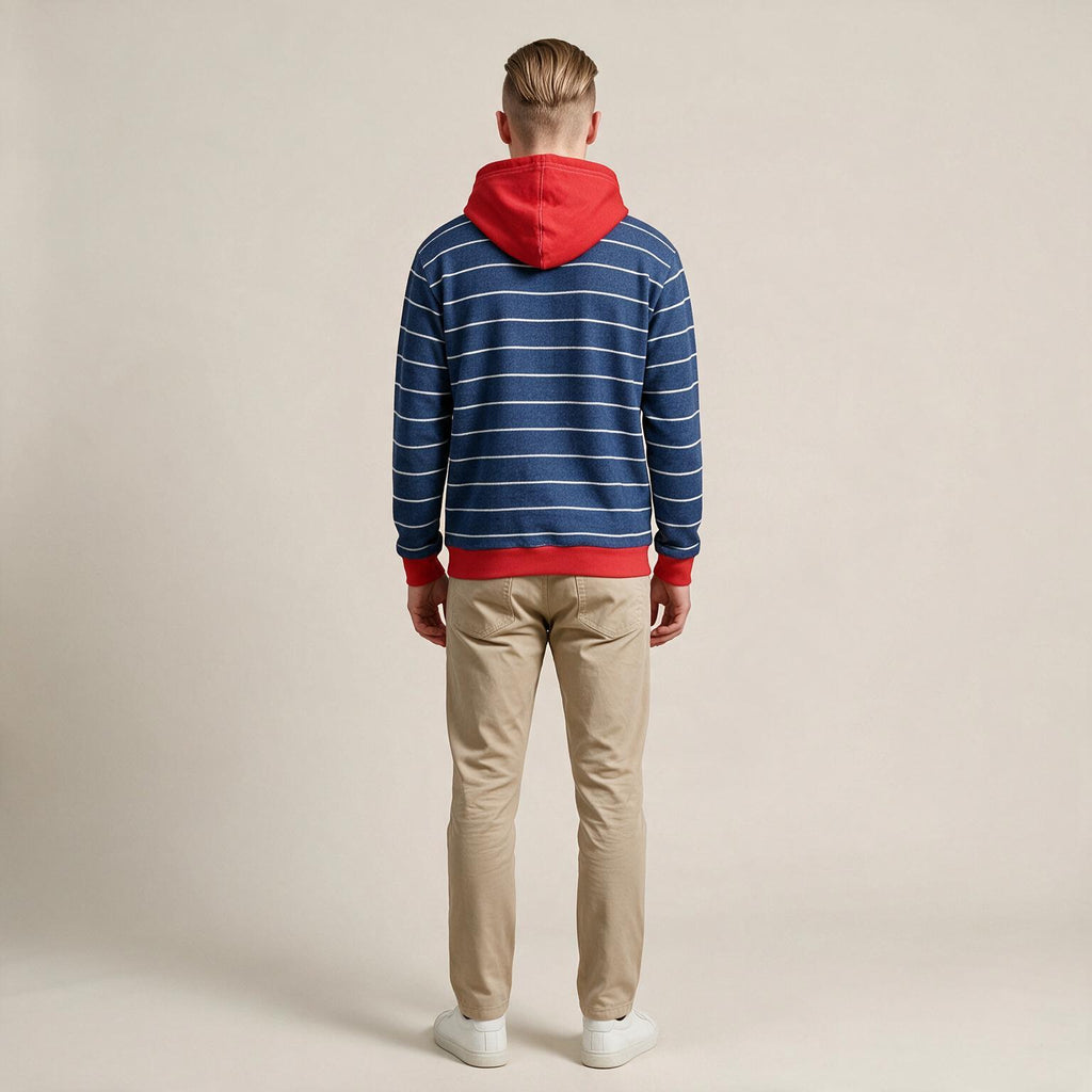 A man viewed from behind wearing a blue and white striped hoodie with a red hood and red trim, paired with beige pants and white sneakers, against a plain beige studio background.