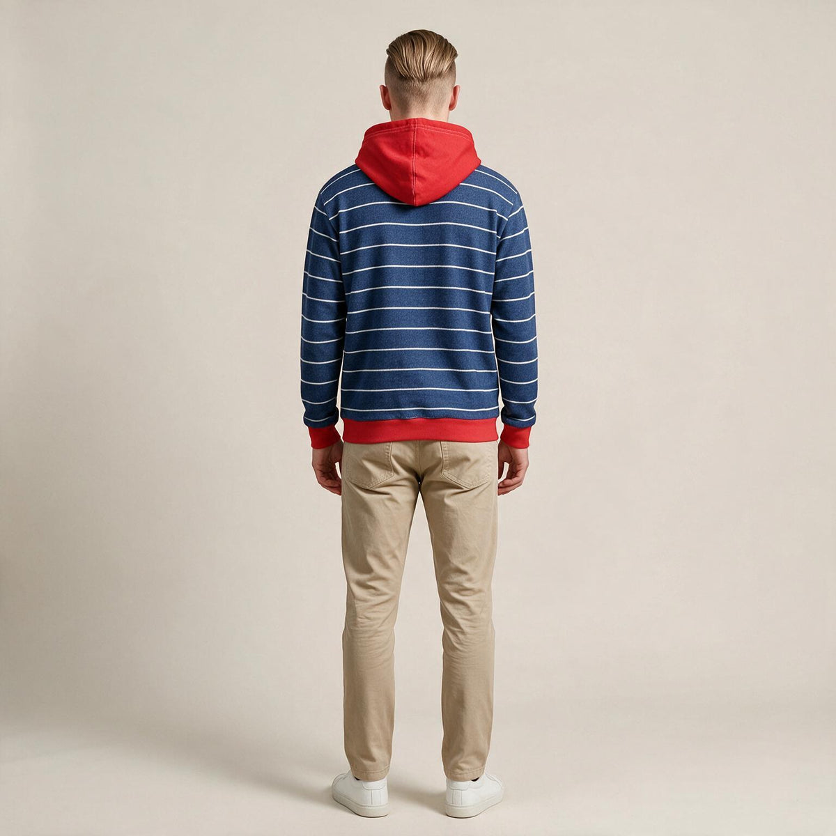 A man viewed from behind wearing a blue and white striped hoodie with a red hood and red trim, paired with beige pants and white sneakers, against a plain beige studio background.
