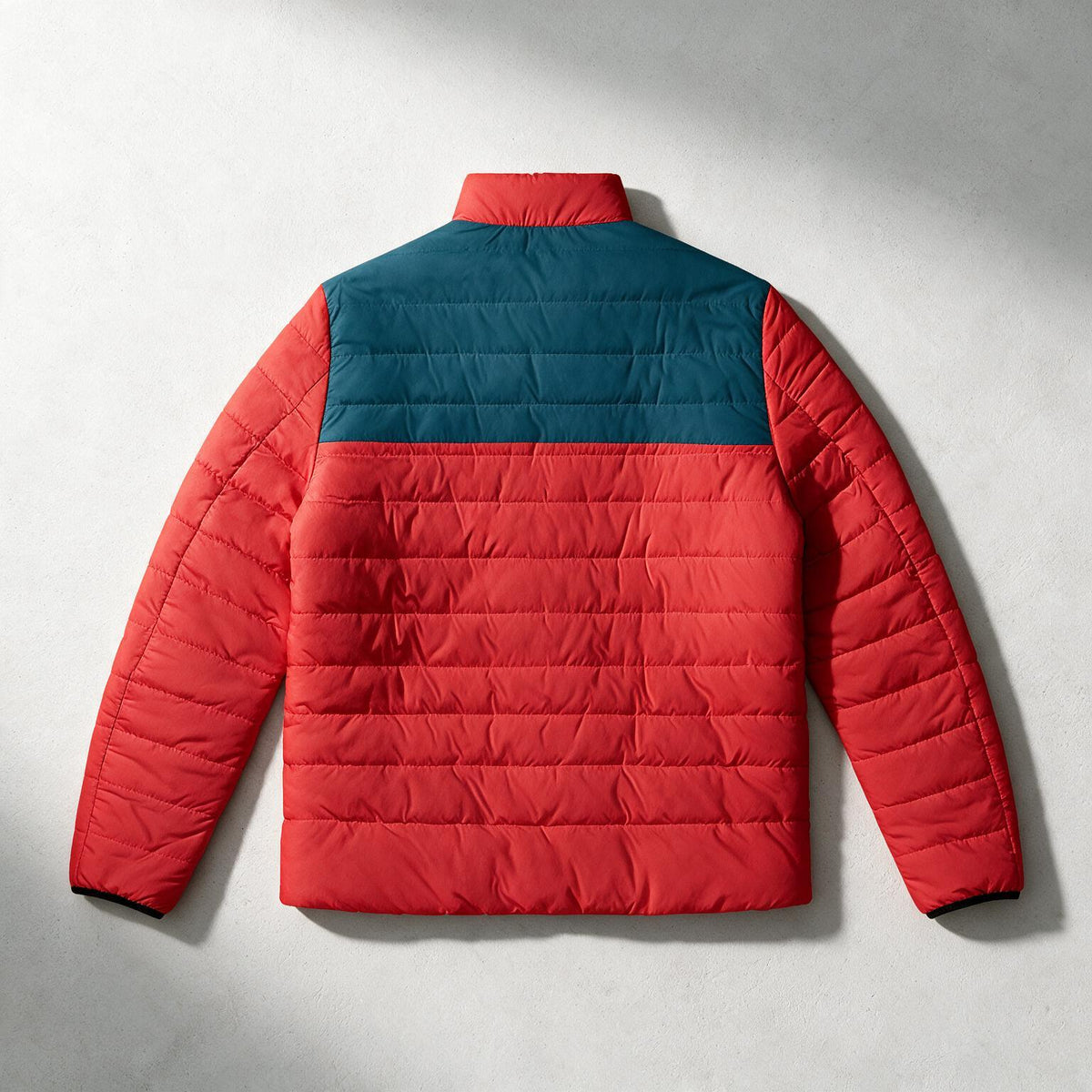 A red and teal color-blocked puffer jacket shown from the back, laid flat on a light gray textured background.