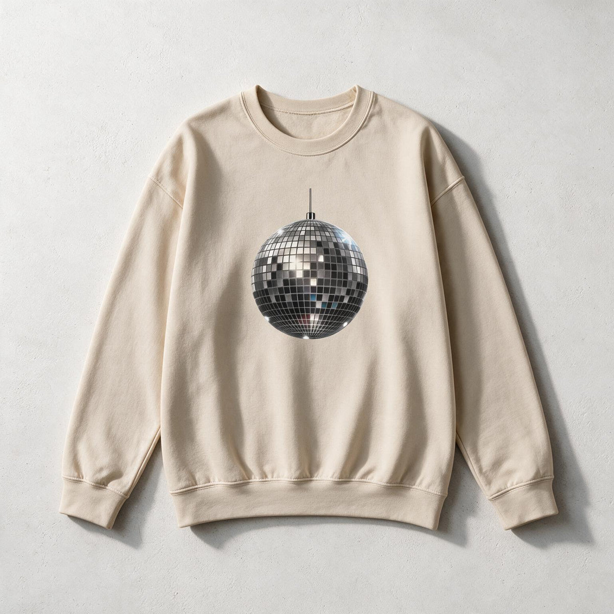 A beige crewneck sweatshirt featuring a graphic print of a silver disco ball in the center, laid flat on a light gray textured background.