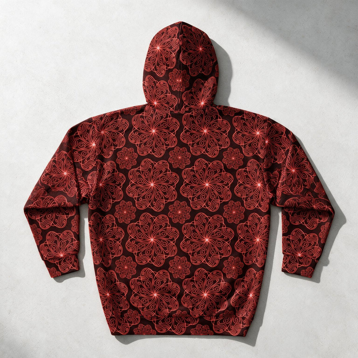 A hooded sweatshirt shown from the back, featuring an all-over red and black floral mandala pattern, laid flat on a light gray textured surface.