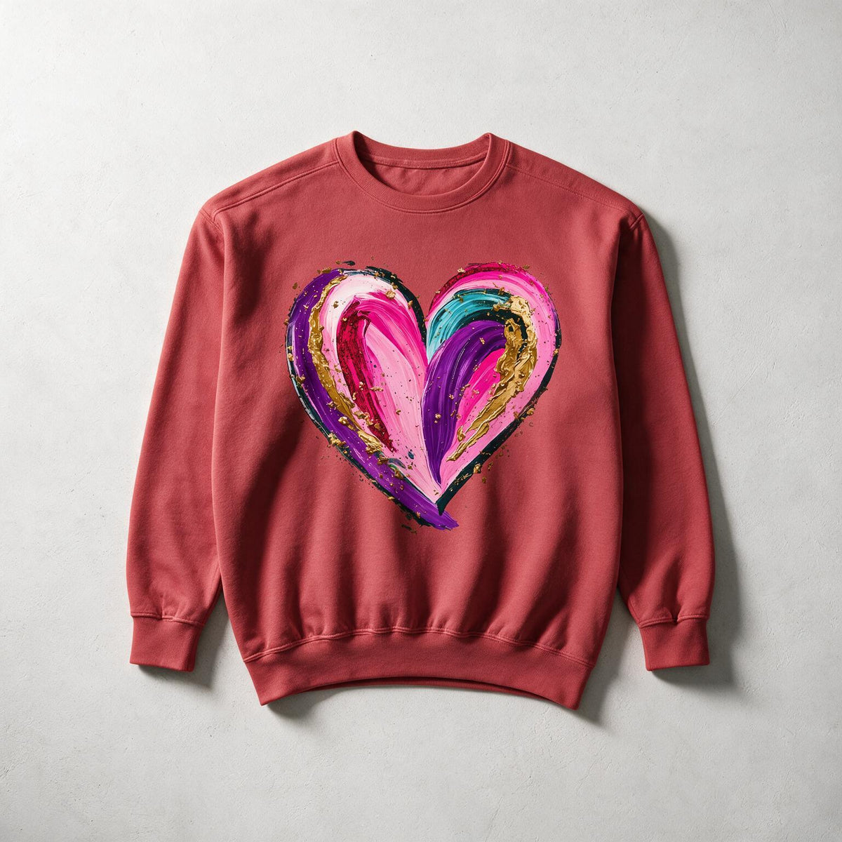 A dusty rose crewneck sweatshirt featuring a large, colorful abstract heart graphic on the chest with pink, purple, teal, and gold paint strokes, laid flat on a light gray textured background.