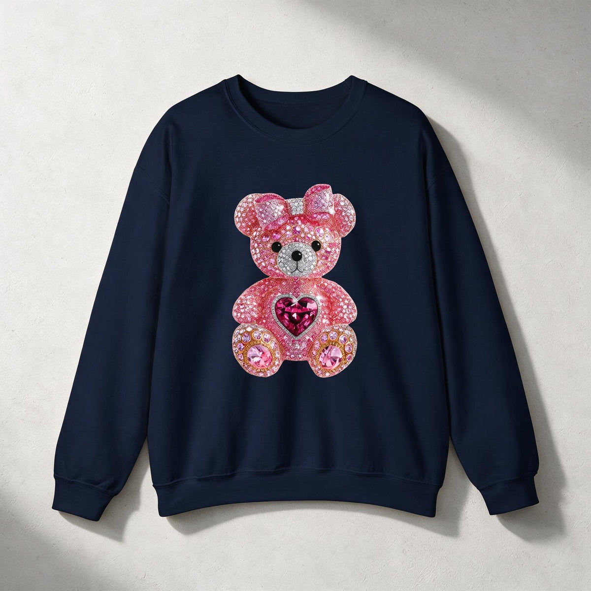 A navy blue crewneck sweatshirt featuring a large pink rhinestone teddy bear graphic on the front, set against a textured light gray background with soft diagonal shadows.