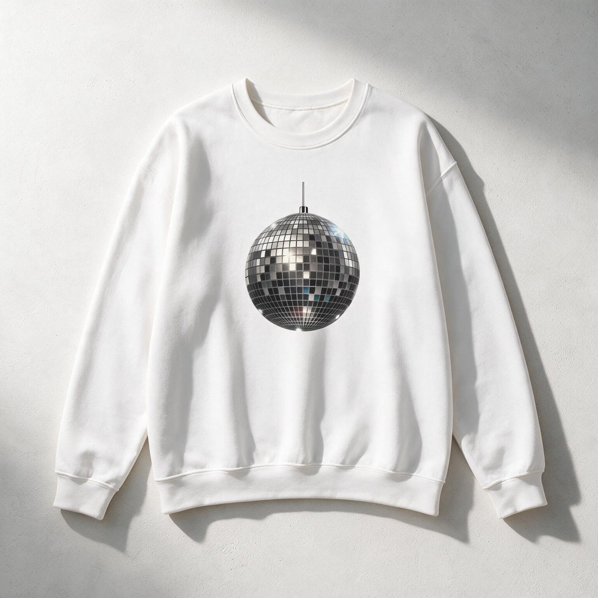 A white crewneck sweatshirt featuring a graphic print of a silver disco ball in the center, laid flat on a light gray textured background with soft shadows.