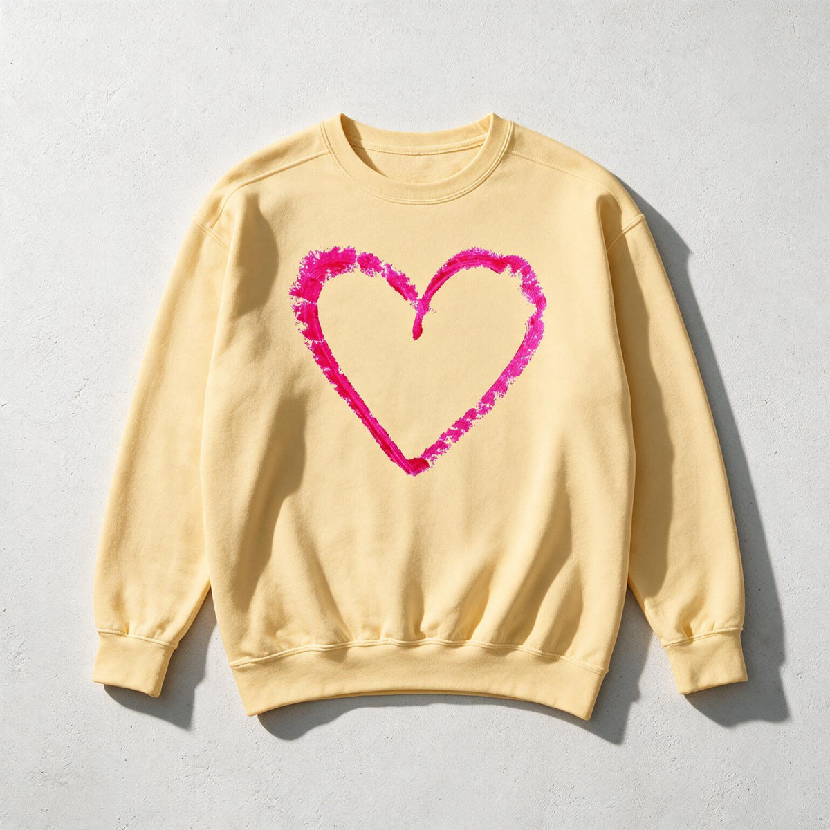 A pale yellow crewneck sweatshirt with a textured, hand-drawn hot pink heart outline on the front, laid flat on a light gray concrete surface.