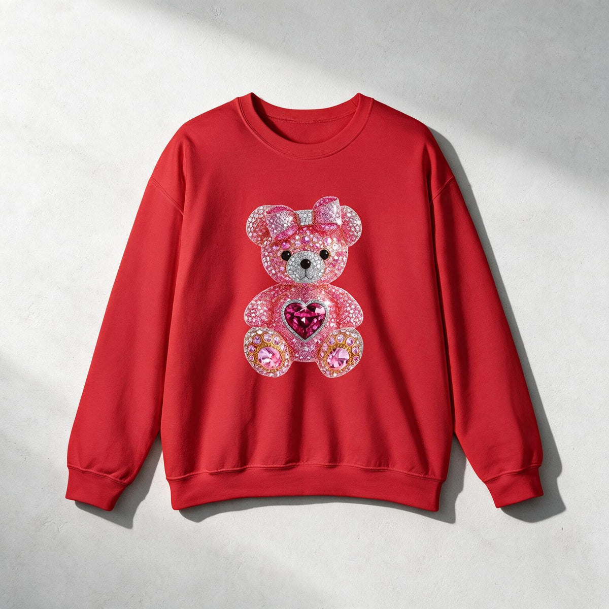 A red crewneck sweatshirt featuring a large, sparkly teddy bear graphic on the front, displayed flat against a textured light gray background with soft diagonal shadows.