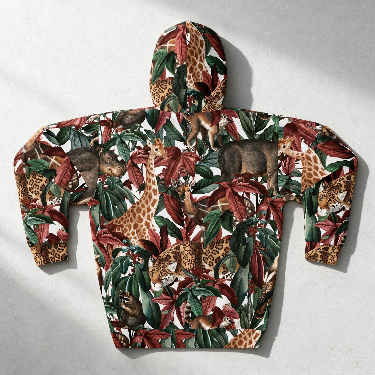 A hooded sweatshirt laid flat on a light gray surface, featuring an all-over safari animal print with giraffes, zebras, leopards, and monkeys among green and red tropical leaves on a white background.