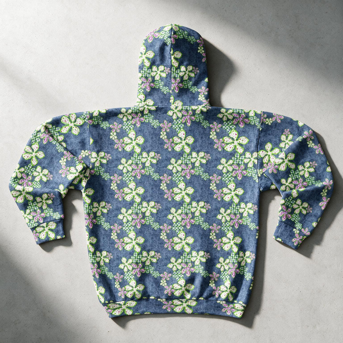 A hooded sweatshirt laid flat on a light gray surface, featuring an all-over floral pattern in light green, lavender, and white on a heathered blue-gray base. The hoodie is photographed from above with soft shadows cast across it.