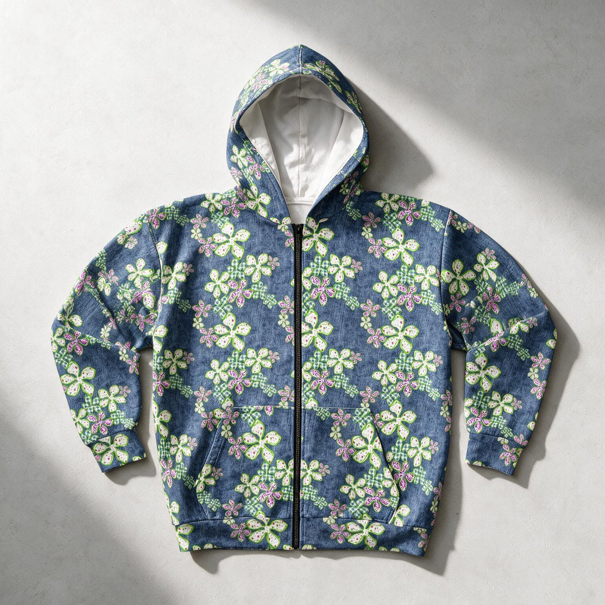A zip-up hoodie with a blue denim-like base and an all-over floral print in green, pink, and yellow, laid flat on a light gray background with soft shadows.