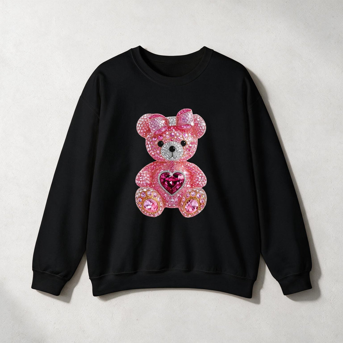 A black crewneck sweatshirt featuring a large, pink rhinestone-embellished teddy bear graphic on the front, set against a light gray textured background.