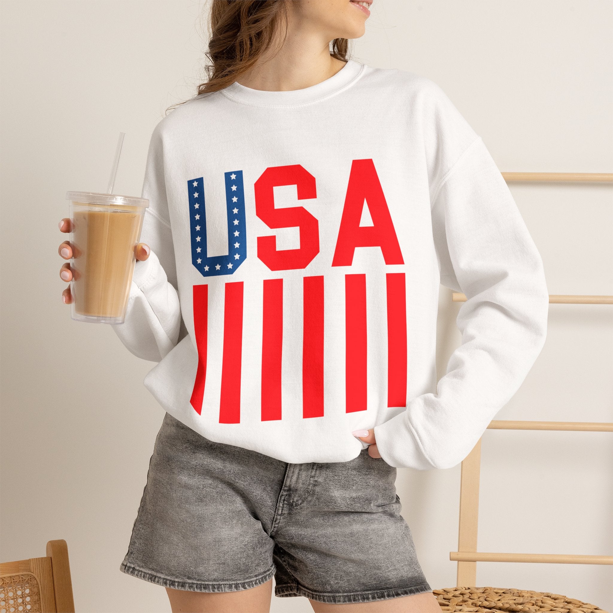 A woman wearing a white crewneck sweatshirt with a patriotic USA graphic in red and blue, paired with gray denim shorts and holding an iced coffee.