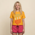 A young woman with blonde hair is smiling and wearing a yellow t-shirt with a distressed red and blue USA flag graphic, paired with hot pink shorts, against a solid beige background.