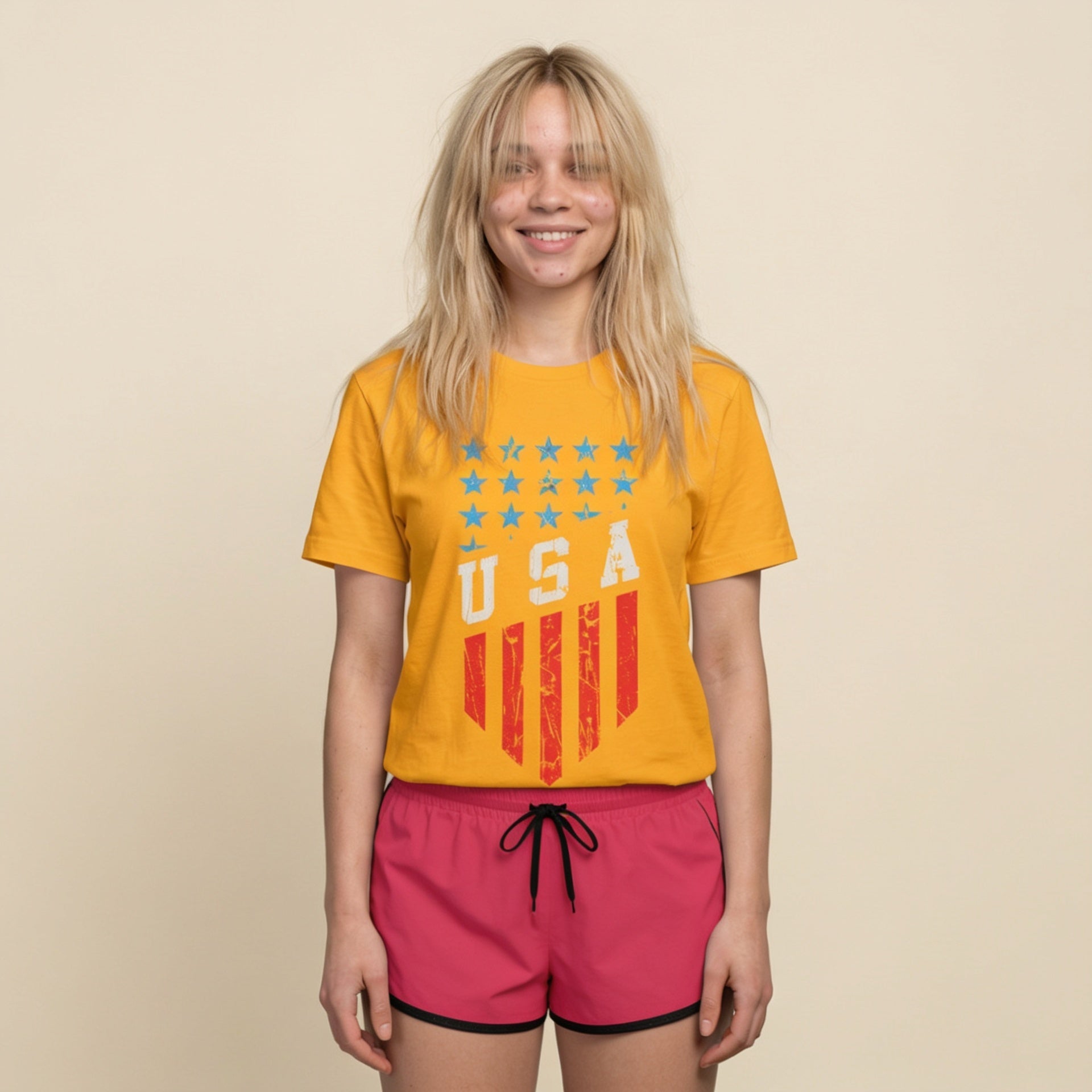 A young woman with blonde hair is smiling and wearing a yellow t-shirt with a distressed red and blue USA flag graphic, paired with hot pink shorts, against a solid beige background.