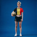 A model stands against a solid blue studio background wearing a cropped t-shirt with black, yellow, and red vertical panels, black denim shorts, and white sneakers, while holding a gold soccer ball.