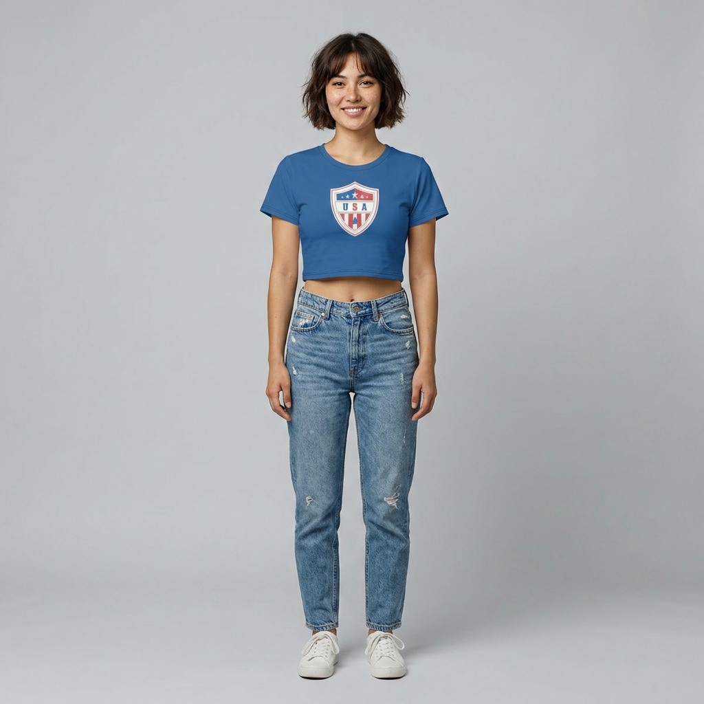 A woman stands against a light gray studio background wearing a blue cropped T-shirt with a red, white, and blue shield graphic on the chest, light-wash blue jeans, and white sneakers.