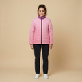 A woman stands against a plain beige studio background wearing a light pink quilted puffer jacket with a contrasting purple collar, black pants, and white sneakers.
