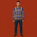 A man stands against a solid orange-red studio background wearing a long-sleeve crewneck sweater with a blue, yellow, and white plaid pattern, dark blue jeans, and dark brown shoes.