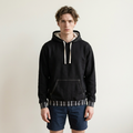 A man wearing a black pullover hoodie with white drawstrings and striped ribbed cuffs and hem, paired with dark denim shorts, standing against a plain light beige background.