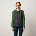 A woman stands against a light textured wall wearing a hooded zip-front jacket with green sleeves and a navy plaid body, paired with dark blue jeans.