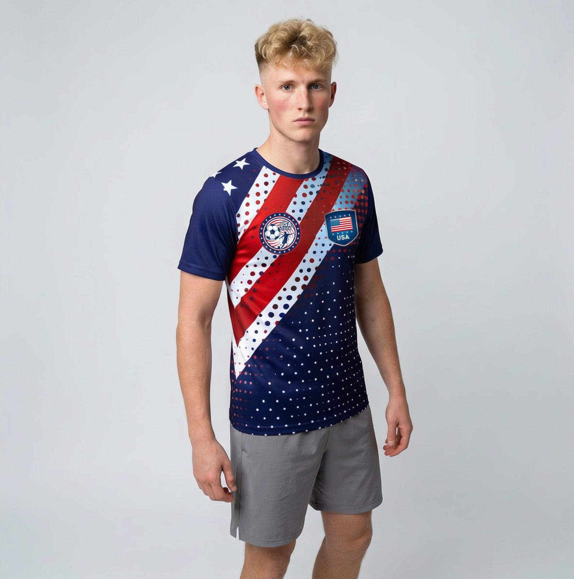 A male model wearing a short-sleeve athletic t-shirt with a red, white, and blue USA soccer theme, paired with gray shorts, against a light gray studio background.
