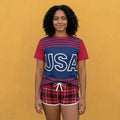 Woman wearing a red and blue striped shirt with 'USA' and plaid shorts against an orange background