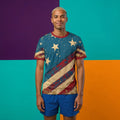 A man wearing a patriotic-themed t-shirt with a distressed American flag pattern and blue shorts, standing against a split purple, teal, and orange background.